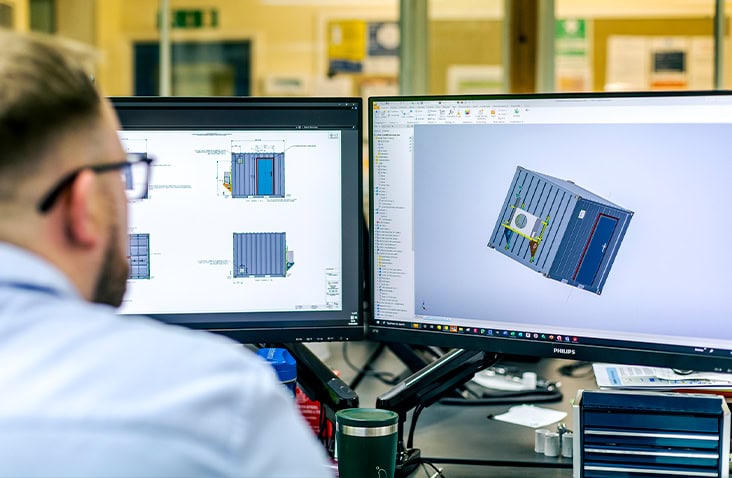 CAD engineer at S Jones Conversions working on dual monitors displaying technical drawings and 3D CAD models of a bespoke shipping container conversion, showing layout planning, ventilation, and equipment integration. 