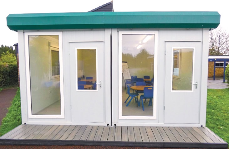 Interior view of a modular classroom at Carrington Junior School, showing a bright teaching space with child-sized tables and chairs visible through glazed doors and windows within a bay modular education unit. 