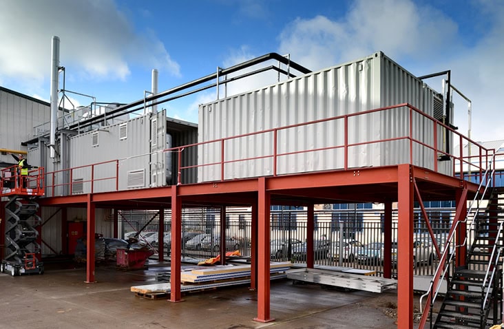 Containerised boiler housing installed on a raised steel platform, showing a white converted shipping container with pipework, ventilation stacks, access stairs and safety railings forming a complete on-site boiler plant solution.