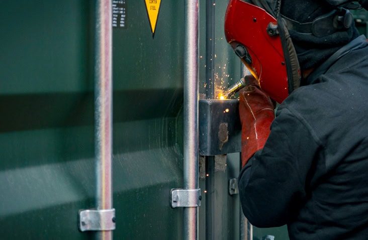 Welder fitting and welding a high-security lock and steel bracket onto a shipping container door as part of a bespoke container security modification