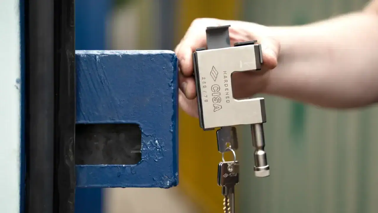Close-up of a person locking a blue shipping container door using a heavy-duty padlock and key, showing container door hardware and security locking mechanism in use.