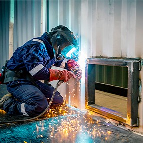 Engineer welding a steel access hatch into the side of a shipping container during structural modification work, demonstrating professional container alteration and fabrication.