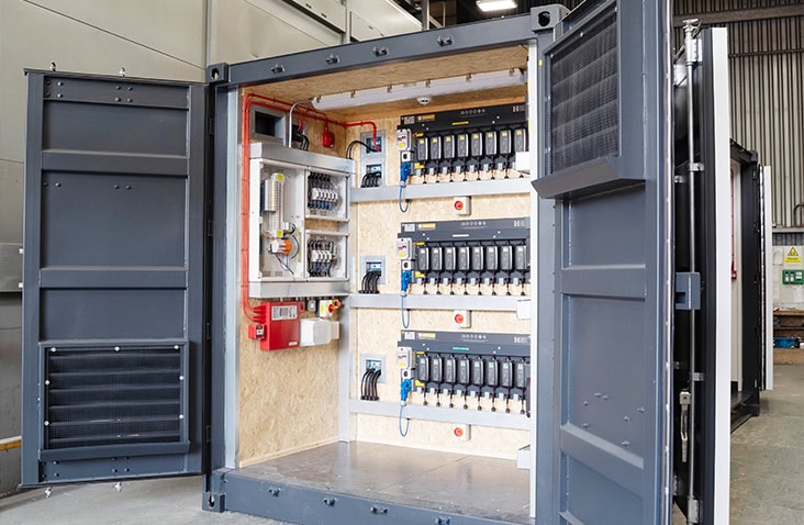 Interior of a containerised electrical control room with open steel doors, displaying mounted control panels, isolators and electrical distribution equipment for battery storage applications. 
