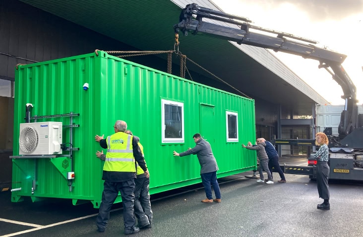 Delivery and positioning of a containerised laboratory using a crane lorry, showing a green converted shipping container with windows and HVAC unit being guided into place by engineers during on-site installation.