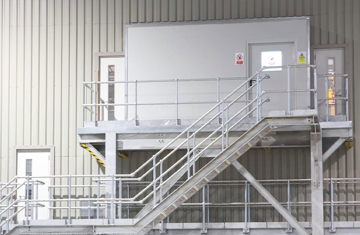 Externally mounted containerised control room conversion with steel access staircase, safety handrails and raised platform, installed inside an industrial facility to provide safe access to an elevated control and monitoring environment.