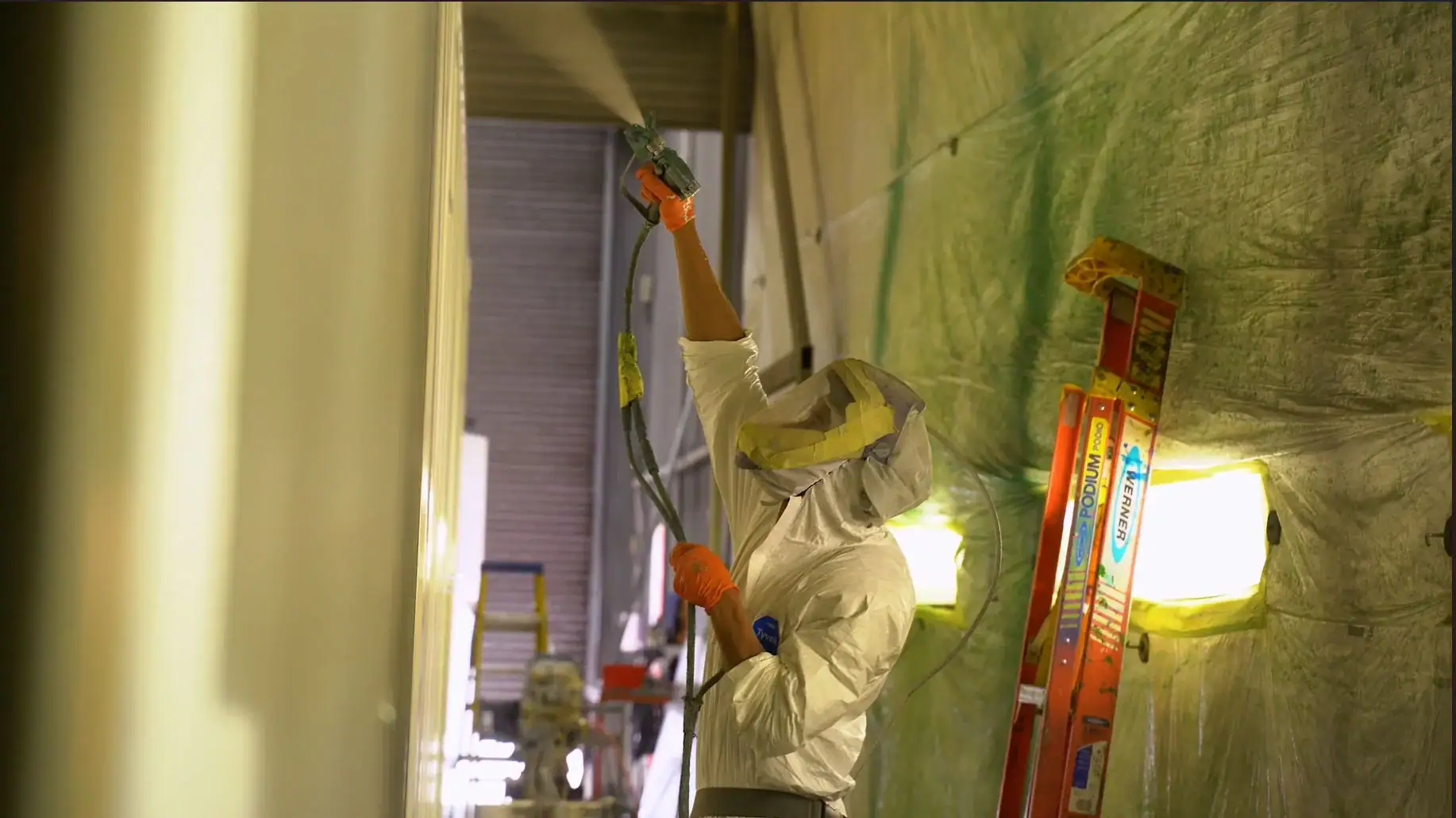 A worker in protective gear, including a full-body suit, gloves, and a hood with a face covering, is spray-painting a large shipping container inside a paint booth. The worker is holding a spray gun high above their head, applying an even coat of paint. The surrounding area is covered in protective plastic sheeting, with bright lights illuminating the workspace. A red ladder and various equipment are visible in the background.