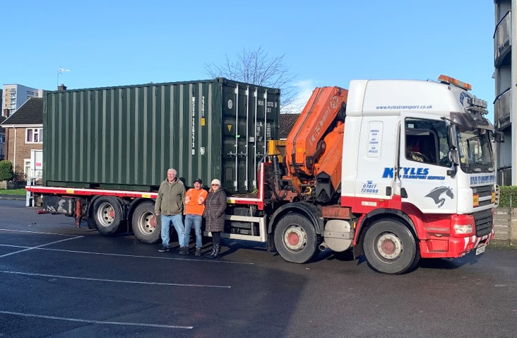 A green 20ft shipping container is being delivered on a flatbed lorry at a residential street, showing crane-assisted container transport for storage or community use. 