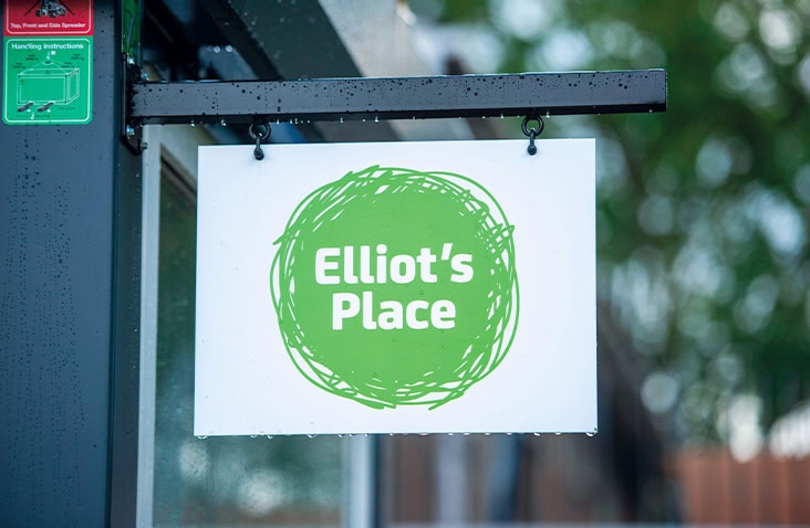 Elliot’s Place signage mounted on a modular building, showing a hanging sign with a green circular logo and white text fixed to the exterior of a modular health sanctuary. 