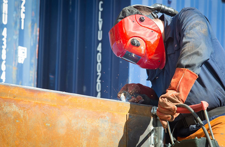 Technician welding a steel shipping container, wearing protective gloves and a red welding mask, with blue containers in the background.