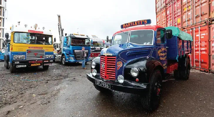 A collection of vintage and classic trucks is parked in an industrial yard with shipping containers in the background. A restored blue and black truck with "Bartlett’s" branding is prominently featured in the foreground, while other colorful classic trucks, including a yellow and blue one, are visible behind it. A man is seen walking among the trucks.