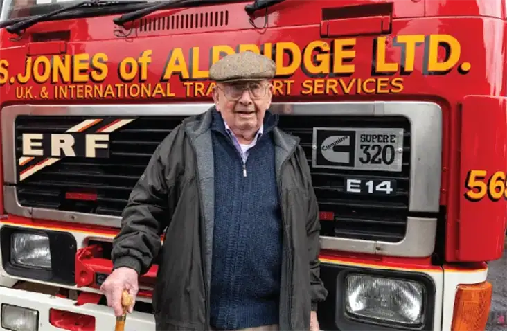 An elderly man wearing a flat cap, glasses, and a dark coat stands in front of a red ERF truck belonging to "S. Jones of Aldridge Ltd." He is holding a walking stick and has a slight smile on his face. The truck has bold yellow lettering and a Cummins "Super 320" badge.