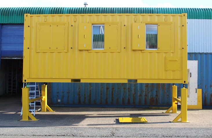 Bright yellow converted shipping container raised on steel support stands, featuring shuttered windows and an access ladder in the background, used as a specialist test and inspection unit at an industrial facility. 