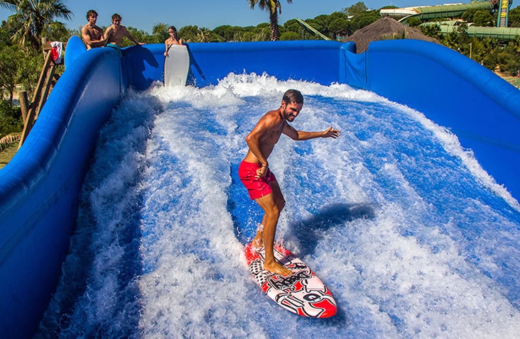 Inflatable flow surfing ride in operation at an outdoor leisure park, showing a surfer riding artificial waves powered by high-capacity water pumps housed in containerised plant equipment. 