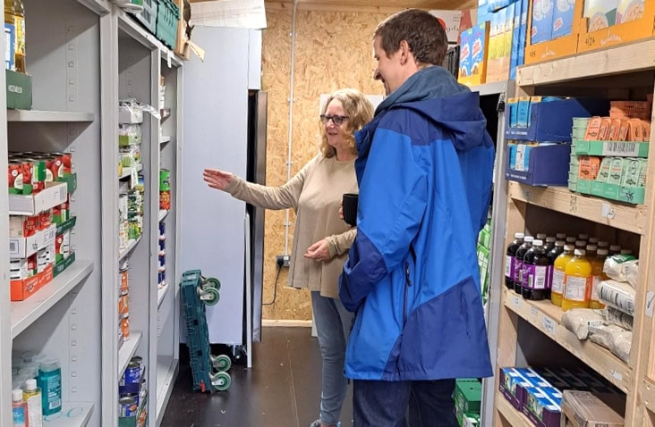Interior of a converted shipping container used as a foodbank store, with shelving stocked with ambient food supplies and volunteers reviewing inventory.