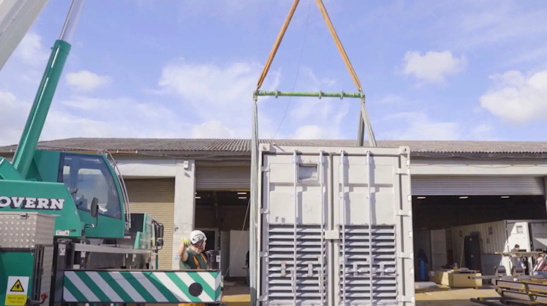 A large industrial crane is lifting a modified shipping container using heavy-duty straps and a spreader bar. A worker in a high-visibility vest and helmet stands nearby, guiding the operation with hand signals. The background features an open workshop with equipment and materials, under a bright blue sky with scattered clouds.