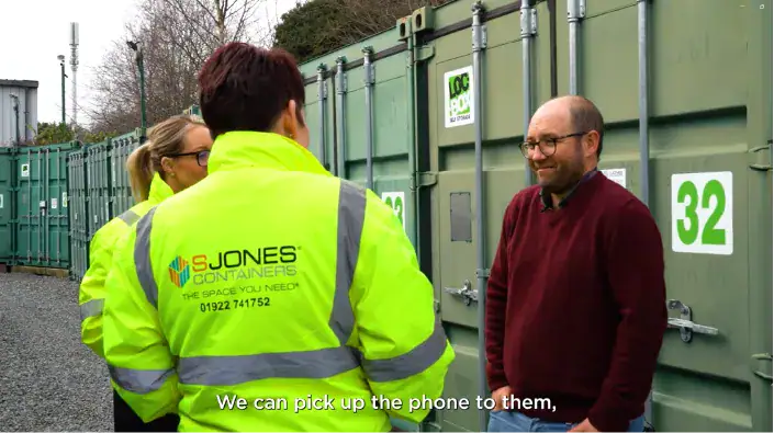 A group of people are having a conversation in front of green shipping containers with numbered labels. Two individuals are wearing high-visibility yellow jackets with 'S Jones Containers' branding, while a man in a burgundy sweater and glasses stands opposite them, smiling. The setting appears to be an outdoor storage facility with gravel ground.