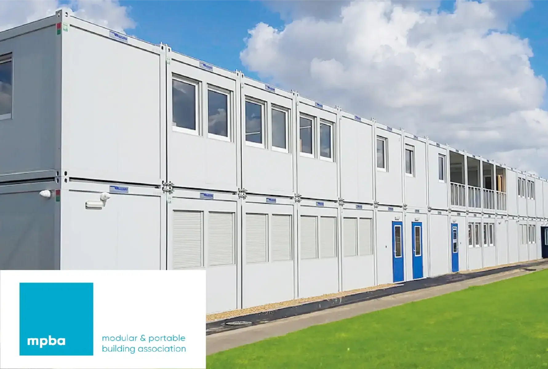 A large two-story modular building made from stacked portable containers, featuring multiple windows, blue entrance doors, and some shuttered sections. The structure is set against a bright blue sky with scattered clouds and surrounded by a well-maintained grassy area. The Modular & Portable Building Association (MPBA) logo is visible in the bottom left corner.