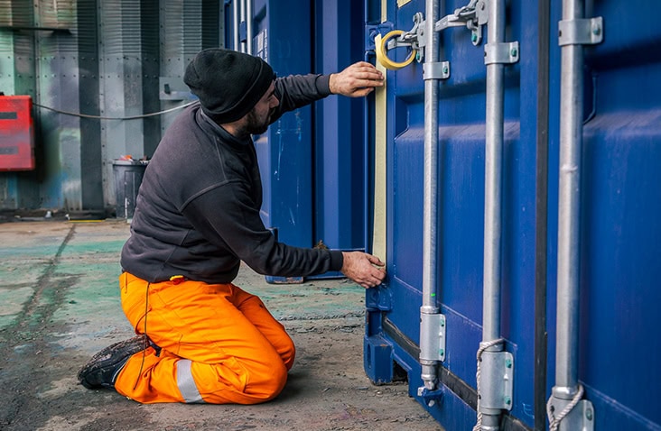 Container conversion technician masking and preparing a blue shipping container door before repainting and surface treatment.