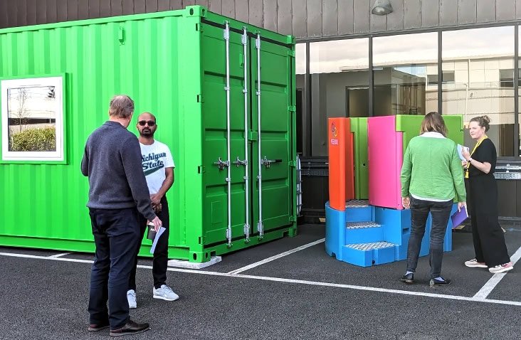 NPK recovery laboratory container installed on a customer site, showing a green converted shipping container alongside external urinal units, with people reviewing the installation and discussing the setup outside the building. 