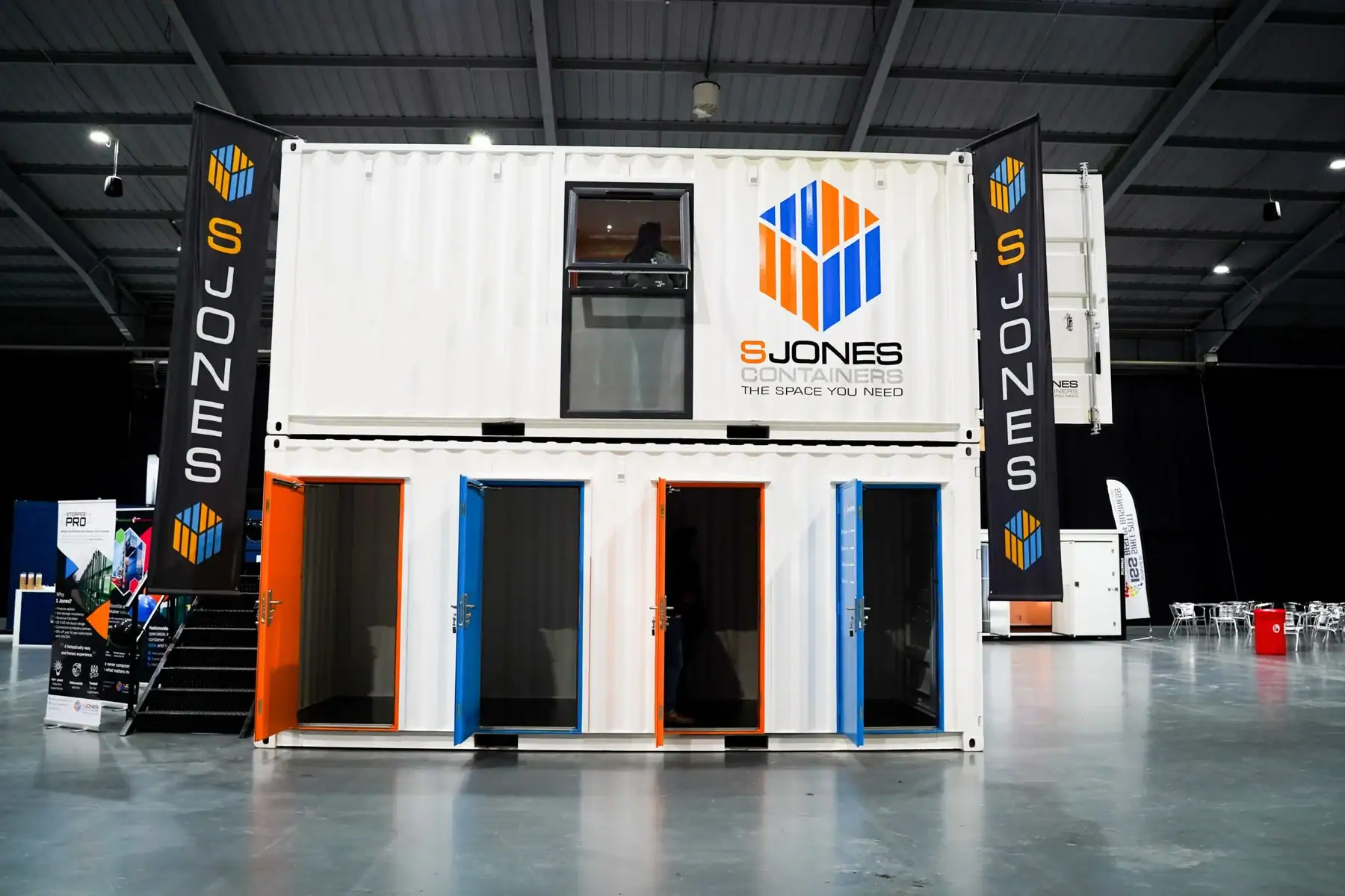 Two-storey S Jones Containers exhibition showbox made from white shipping containers, featuring branded signage, external staircase, and multiple coloured entrance doors, displayed indoors at a trade exhibition hall.