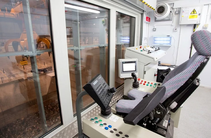 Interior of a containerised control room conversion featuring an operator control chair, joystick controls, touch screen monitors and control panels, with large observation windows overlooking an industrial processing area, designed for remote plant and machinery operation. 