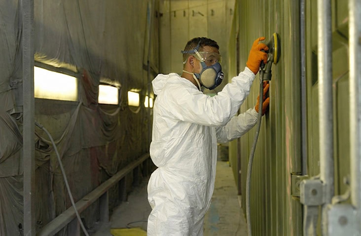 A person wearing a white protective suit, orange gloves, and a respirator is sanding the surface of a green shipping container inside a paint booth.