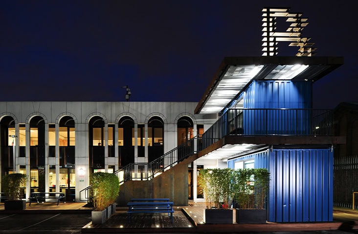 Pop-up containerised café at Battersea Studios photographed at night, showing stacked blue shipping containers with external stair access, terrace seating, architectural lighting and landscaped surroundings in an urban courtyard. 