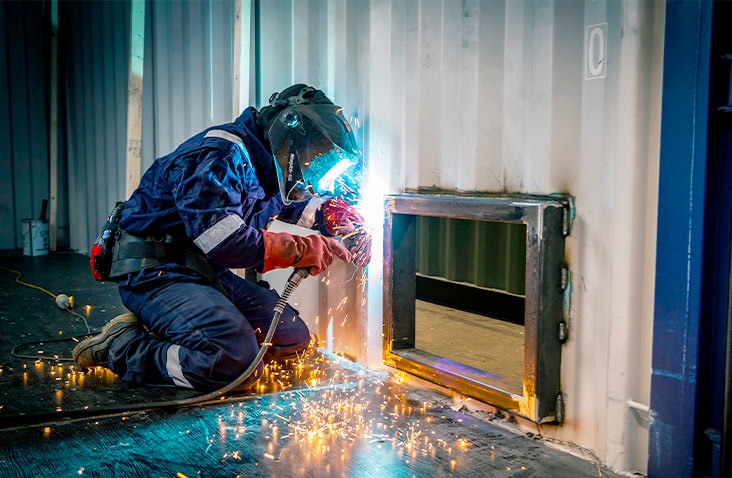 A welder in protective gear kneels beside a shipping container wall, using a welding torch to cut and shape a framed opening. Bright sparks scatter across the floor as the work is carried out inside a workshop environment.