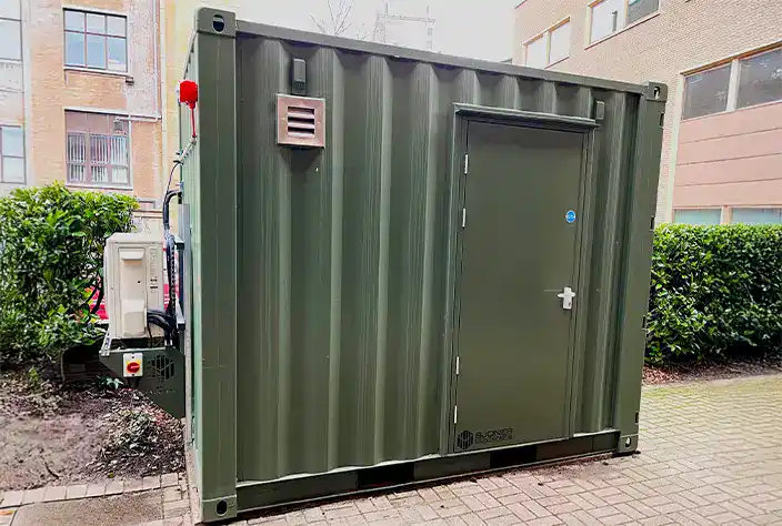 A green converted shipping container unit with a solid door, wall-mounted air conditioning unit, and ventilation grille, situated on a paved area next to a building and hedges.