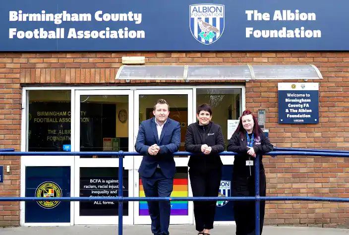 Three people stand smiling outside the Birmingham County Football Association and The Albion Foundation office, which has a blue sign with logos above the entrance and a rainbow flag displayed in the window.