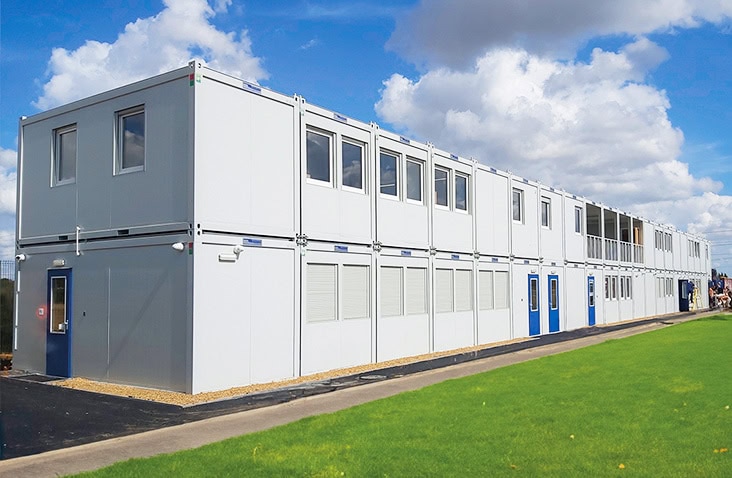 Two-storey modular school building with white panel exterior, multiple windows and blue doors, installed alongside a grass field.