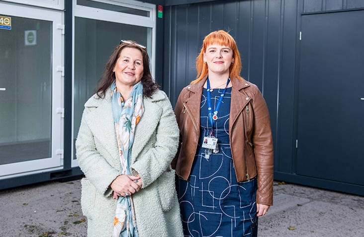 Two women standing outside a completed modular unit at Elliot’s Place, showing the exterior of a modular building with glazed entrance doors and modern cladding. 