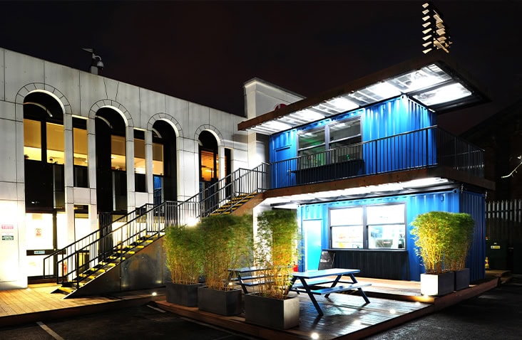 Two-storey shipping container café conversion at Battersea Studios, featuring stacked containers, external steel staircase, rooftop terrace, glazed service hatch and outdoor seating, photographed at night.