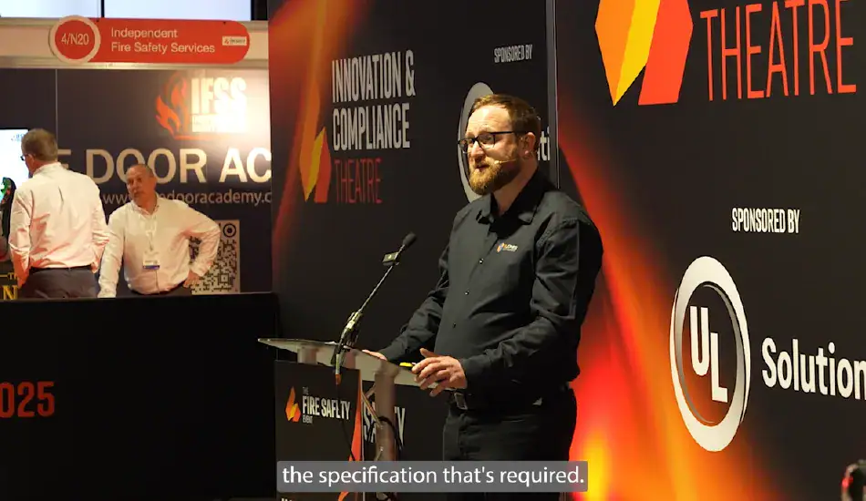Speaker presenting at a lectern on a theatre stage at the Fire Safety Event 2025, with event branding and sponsor logos visible on the backdrop, representing industry talks and presentations within the fire safety and compliance sector.