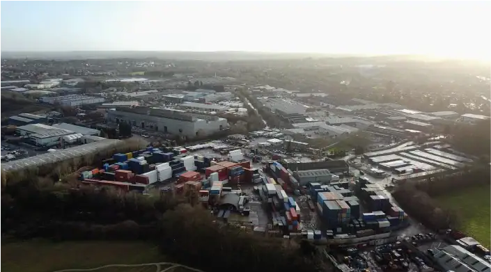 An aerial view of an industrial area with large warehouses, buildings, and a shipping container yard filled with colorful stacked containers. The surrounding landscape includes trees, roads, and an open field.