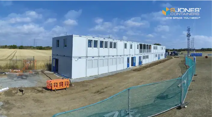 A large modular container building painted white and arranged in a double-stacked structure is under construction on a fenced-off site. The S Jones Containers logo and tagline 'The Space You Need' are visible in the top right corner of the image.