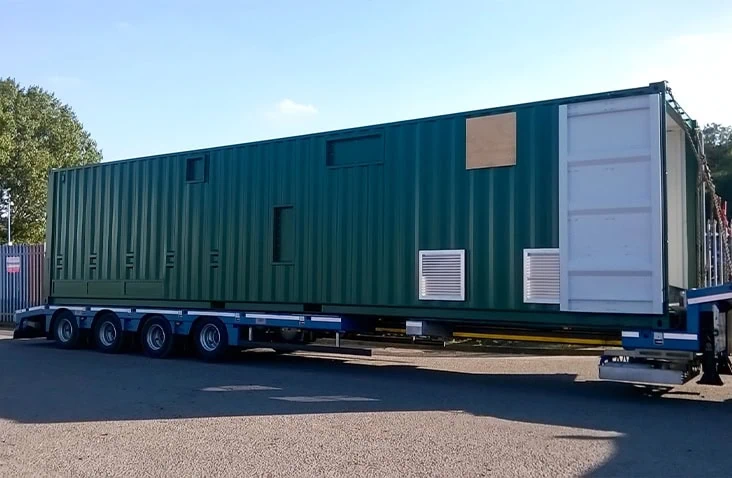 40ft shipping container converted into a water treatment plant during delivery, showing a green containerised treatment unit with ventilation louvres and access points transported on a flatbed trailer.